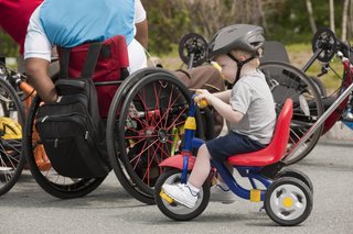 Ein kleiner Junge sitzt auf einem Dreirad, er trägt einen Fahrradhelm. Hinter ihm sieht man einen Erwachsenen, der im Rollstuhl sitzt. 