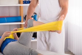 Ein Physiotherapeut macht Übungen mit einer Patientin, die auf einer Liege liegt, dabei nutzt er ein gelbe Thera-Band.