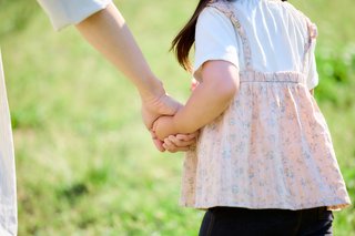 Ein kleines Mädchen an der Hand seiner Mutter, sie gehen gemeinsam über eine Wiese und sind nur von hinten zu sehen.