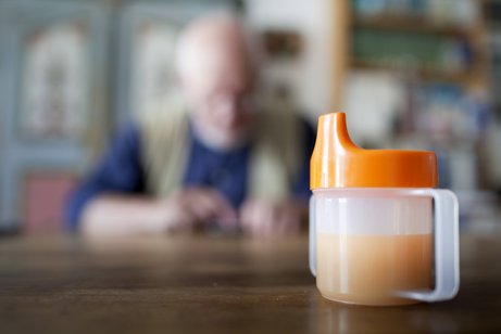 Pflege zu Hause Eine Schnabeltasse steht auf einem Tisch. Im Hintergrund sieht man unscharf einen alten Mann am Tisch sitzen.