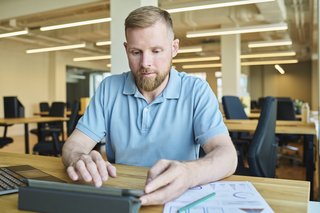 Ein Mann mittleren Alters sitzt an seinem Schreibtisch in einem Büro, er arbeitet an einem Tablet.