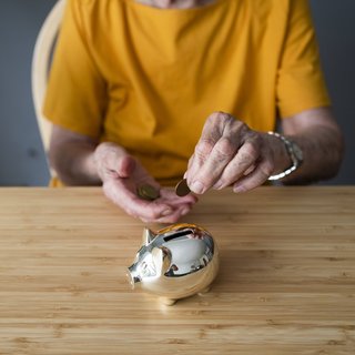 Eine ältere Frau sitzt an einem Tisch, steckt Münzen in ein goldenes Sparschwein.