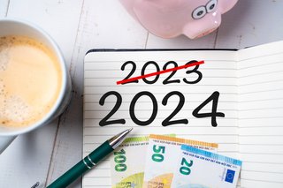 Ein Notizblock mit der Zahl 2024 und einer durchgestrichenen Zahl 2023 auf einem Schreibtisch zusammen mit einem Sparschwein und Euroscheinen. 