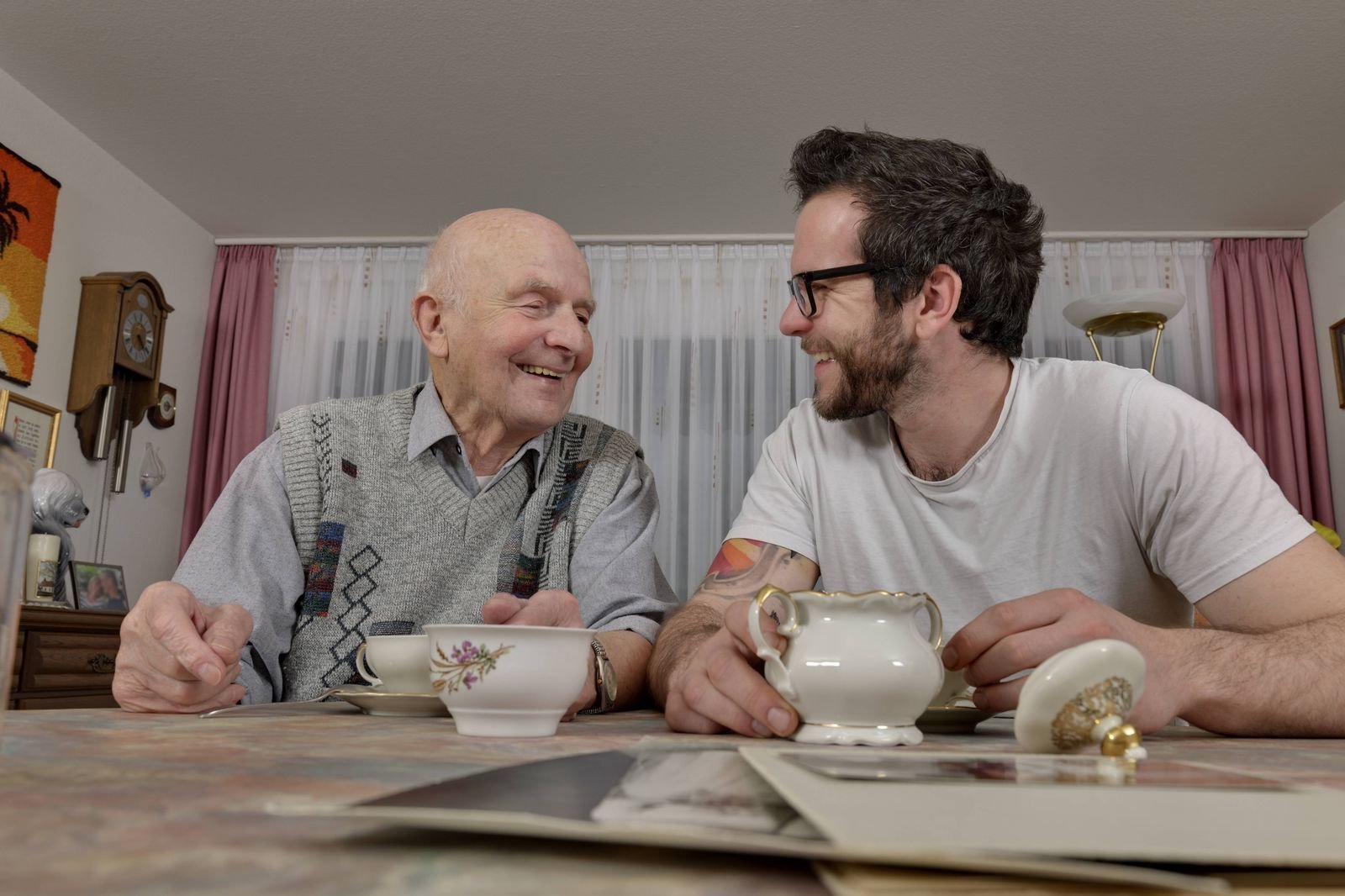  Älterer und junger Mann trinken gemeinsam Tee und lachen miteinander als Symbol dafür, dass Modell wie "Wohnen für Hilfe" funktionieren können.