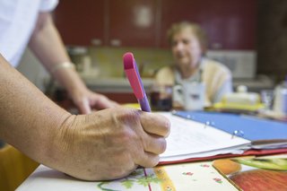 Ältere Frau am Küchentisch. Im Vordergrund Hand einer Frau, sie macht gerade Notizen