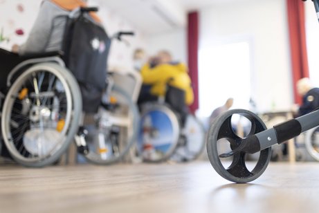 Pflegeheim Blick in einen Raum in einem Pflegeheim, mehrere Menschen mit Rollstühlen sitzen an Tischen, im Vordergrund sieht man ein Rad eines Rollators