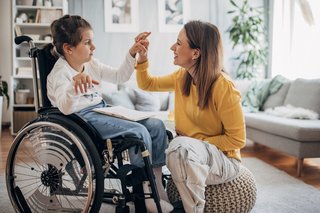 Mutter kniet vor ihrer Tochter mit einer Behinderung, die im Rollstuhl sitzt, und hält ihre Hand