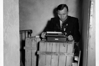 Auf dem Foto sieht man einen ehrenamtlichen VdK‐Mitarbeiter aus der Nachkriegszeit in schwarz-weiß. Er steht vor einer Schreibmaschine, die auf einer Holzkiste steht. 