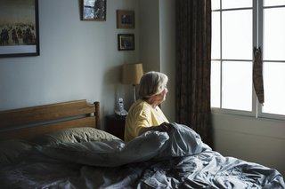 Eine ältere Frau sitzt auf der Kante ihres Bettes und schaut aus dem Fenster