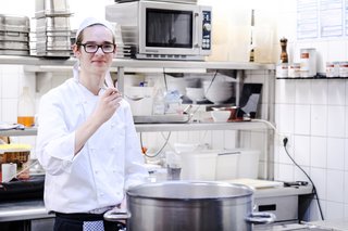 Ein junger auszubildender Koch steht in einer Restaurantküche hinter einem großen Kochtopf und hält einen Löffel zum Kosten vor sich.