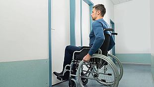 Symbolbild: Junger Mann im Rollstuhl steht vor einer verschlossenen Tür.
