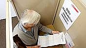 Eine ältere Frau in einer Wahlkabine, sie hält einen Stimmzettel in der Hand.