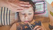 ein Kind hält ein Fieberthermometer am Mund, eine Hand liegt auf der Stirn
