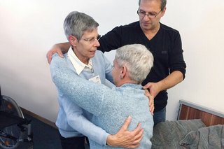 Eine Frau hebt eine andere Frau aus dem Bett, ein Mann gibt Hilfestellung