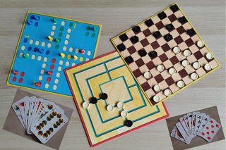 Bild zeigt drei Brettspiele mit Spielsteinen (Mensch ärger dich nicht, Damespielbrett und Mühlenspielbrett) und zwei Kartenspielblätter Skat und Schafkopf auf einem Tisch