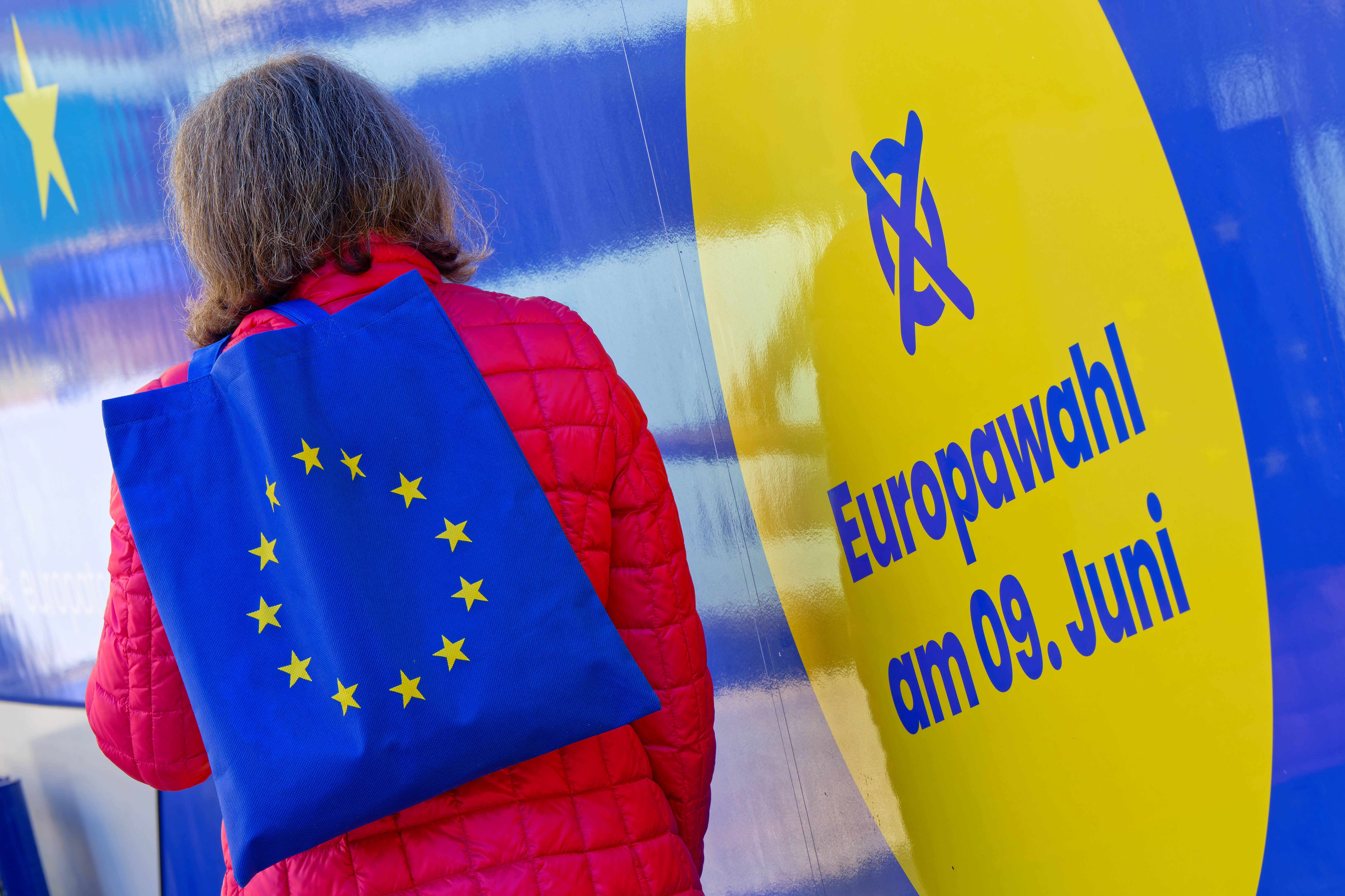Eine Frau mit einer Tasche, die als Aufdruck die Flagge der Europäischen Union hat, steht vor einem blau-gelben Plakat mit der Aufschrift "Euriopawahl am 9. Juni". 