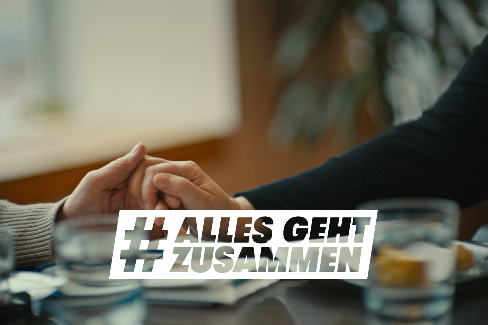 Zwei Menschen halten sich tröstend und unterstützend an der Hand, sie sitzen an einem Tisch. Im Vordergrund das Logo Alles geht zusammen