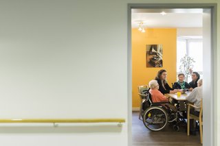 Blick in den Flur eines Pflegeheims, durch eine offene Tür sieht man eine Gruppe Pflegebedürftiger und eine Pflegerin an einem Tisch sitzen.