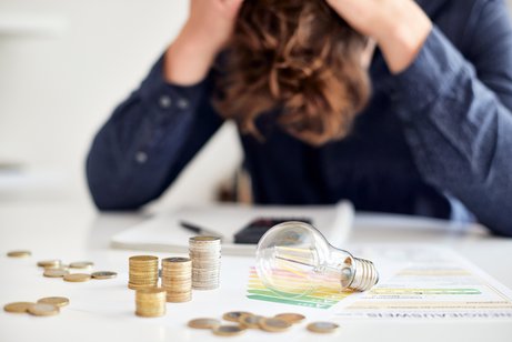 Symbolfoto für Energieschulden: Ein junger Mann sitzt an einem Tisch, auf dem Münzen, Schreiben und eine Glühbirne liegen. Er hat den Kopf gesenkt, rauft sich die Haare.