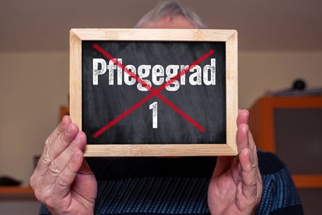 Pflegegrad 1 Fotomontage: Ein älterer Mann hält ein Schild mit dem durchgestrichenen Schriftzug: Pflegegrad 1. Das Symbolbild steht für die Abschaffung des Pflegegrads 1.
