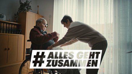 Eine ältere Frau legt einem älteren Mann, der im Rollstuhl sitzt, eine Decke über die Beine. Darüber das Logo der Aktion Alles geht zusammen.