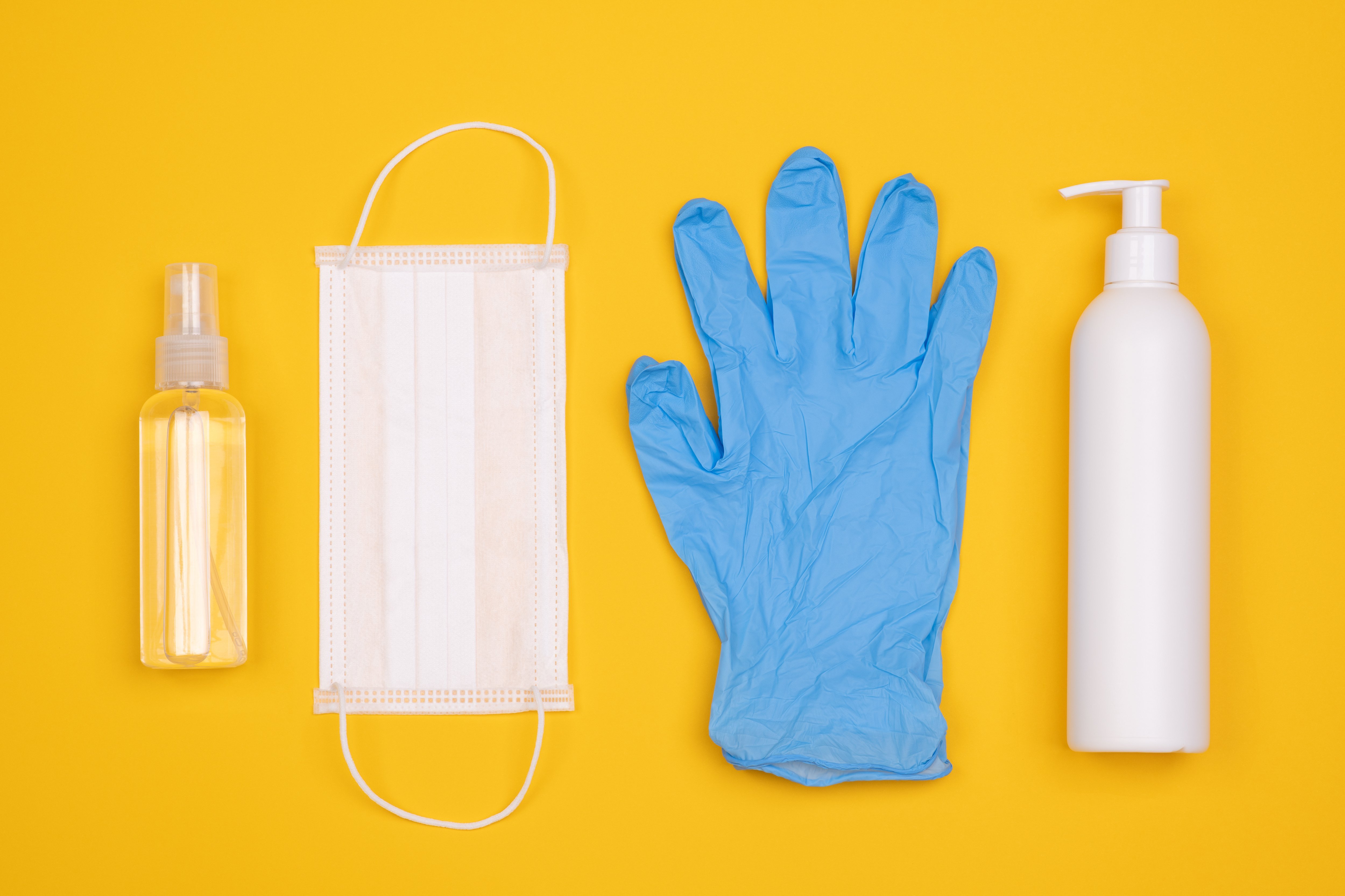 Pflegehilfsmittel zum Verbrauch Eine Flasche mit Desinfektionsmittel, ein Mundschutz, ein blauer Gummihandschuh uind eine Flasche mit einer Lotion nebeneinander vor gelbem Hintergrund