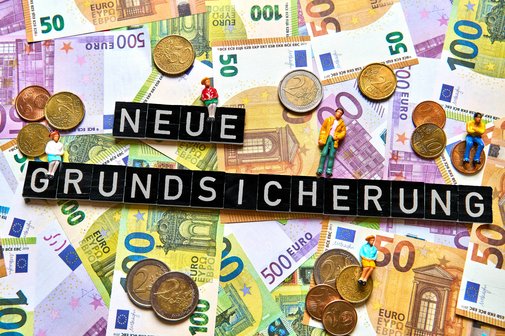 Geldscheine und Münzen, darauf bilden Buchstabenwürfel die Worte "Neue Grundsicherung"