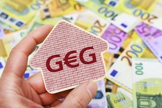 Gebäudeenergiegesetz Eine Hand hält ein Schild mit der Aufschrift "GEG", da E ist ein Eurozeichen. Dahinter liegen viele Euro-Scheine. |