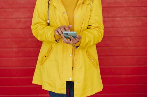 Frau tippt auf ihrem Smartphone Eine Frau in einem gelben Regelmantel steht vor einer roten Steinmauer, tippt etwas in ihr Smartphone.