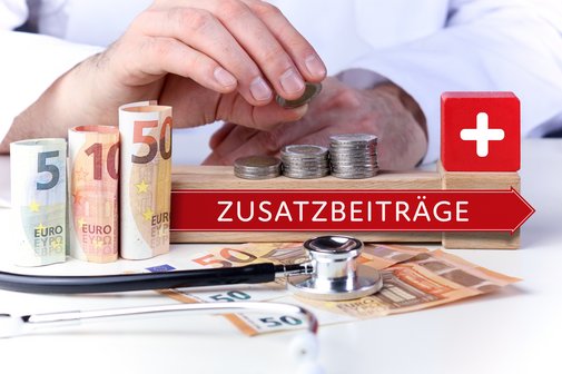 H&auml;nde legen M&uuml;nzen auf gestapelte Euro-M&uuml;nzen und Geldscheine; daneben ein roter Pfeil mit der Aufschrift &bdquo;Zusatzbeitr&auml;ge&ldquo;, ein roter W&uuml;rfel mit wei&szlig;em Kreuz und ein Stethoskop &ndash; Symbolbild f&uuml;r steigende Zusatzbeitr&auml;ge im Gesundheitswesen.