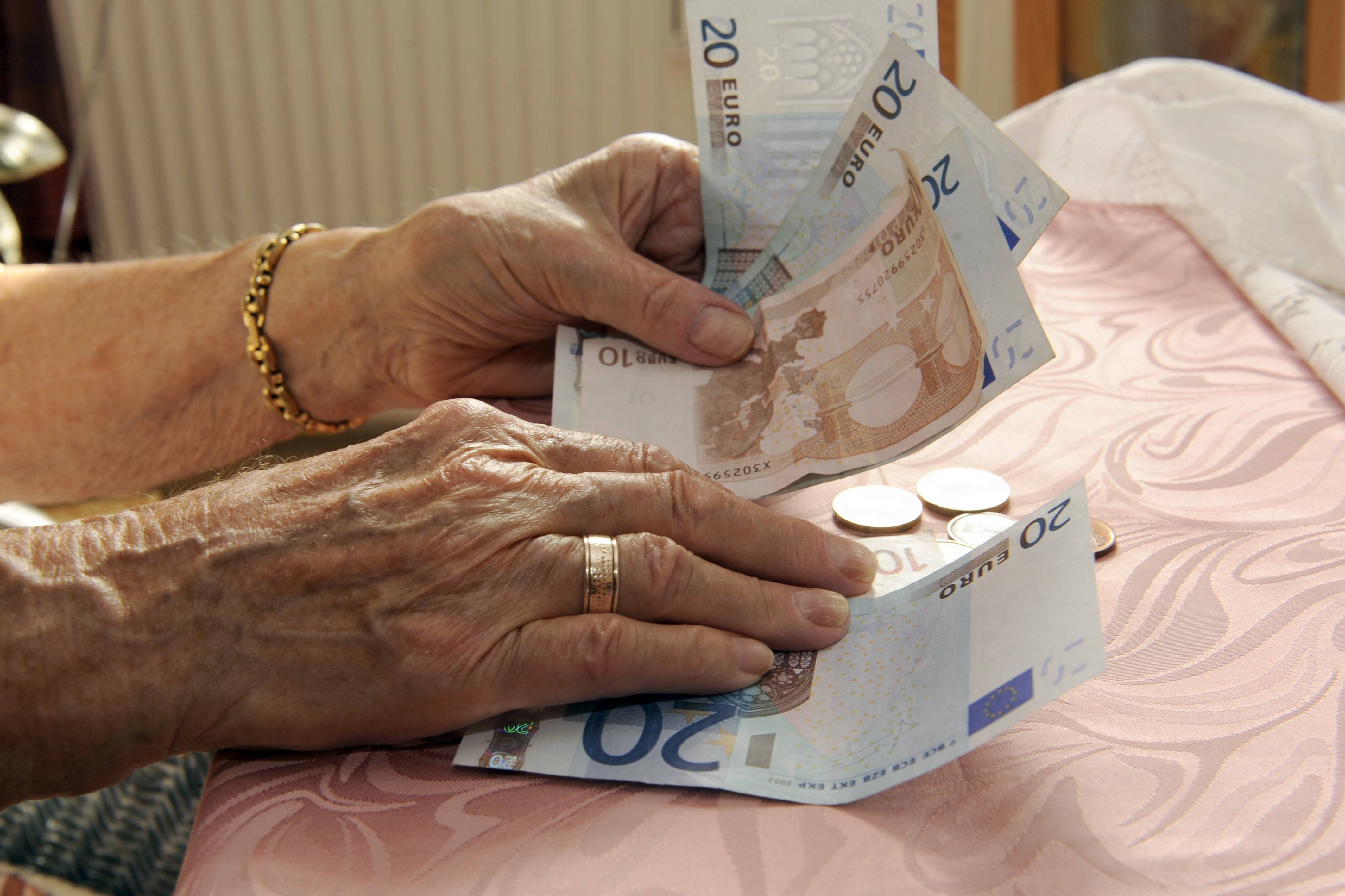 Händer einer alten Frau, die an einem Tisch sitzt, eventuell in einem Pflegeheim. Sie hält Geldscheine, auf dem Tisch liegen Münzen. 
