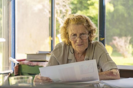 Abrechnung des Pflegedienstes Konzentrierte ältere Frau mit Brille, die an ihrem Schreibtisch in einem sonnendurchfluteten Raum mit Blick ins Grüne sitzt und Papierkram durchgeht, möglicherweise Rechnungen oder medizinische Unterlagen
