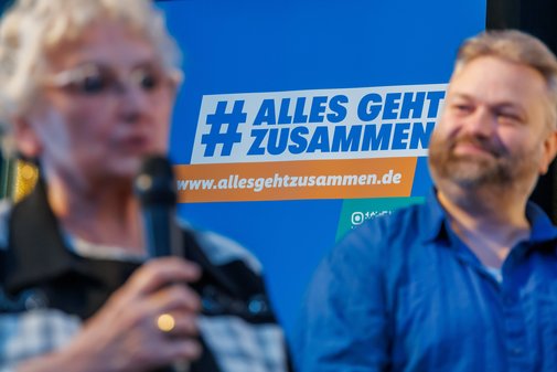 Zwei ehrenamtlich Engagierte - eine Frau und ein Mann - stehen vor einem Roll-up mit dem Aktionslogo von Alles geht zusammen, die Frau spricht gerade ins Mikrofon.