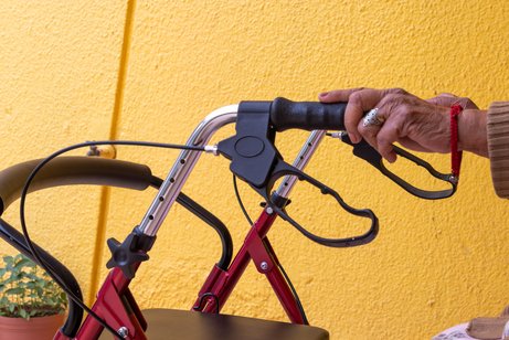 Pflege im Alter Hände einer alten Frau halten die Griffe eines Rollators. Im Hintergrund eine gelb gestrichene Wand.