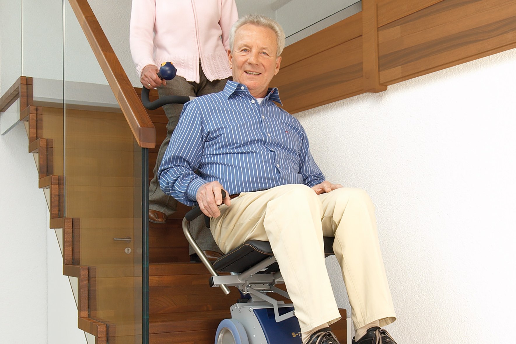 Treppensteighilfe Eine ältere Frau schiebt ihren Mann mithilfe einer Treppensteighilfe - ein Hilfsmittel ähnlich einem Rollstuhl oder Krankentransportstuhl - eine Treppe hinunter.