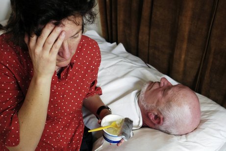Eine Frau sitzt am Bett eines älteren Mannes, der im Bett liegt. Sie hält einen geöffneten Joghurt mit einem Löffel darin in der Hand, fasst sich mit einem Ausdruck der Erschöpfung an die Stirn.