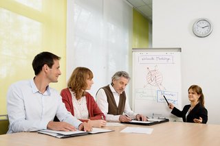 Zwei Männer und zwei Frauen im Büro, eine der beiden Frauen zeigt etwas an einem Flipchart. 