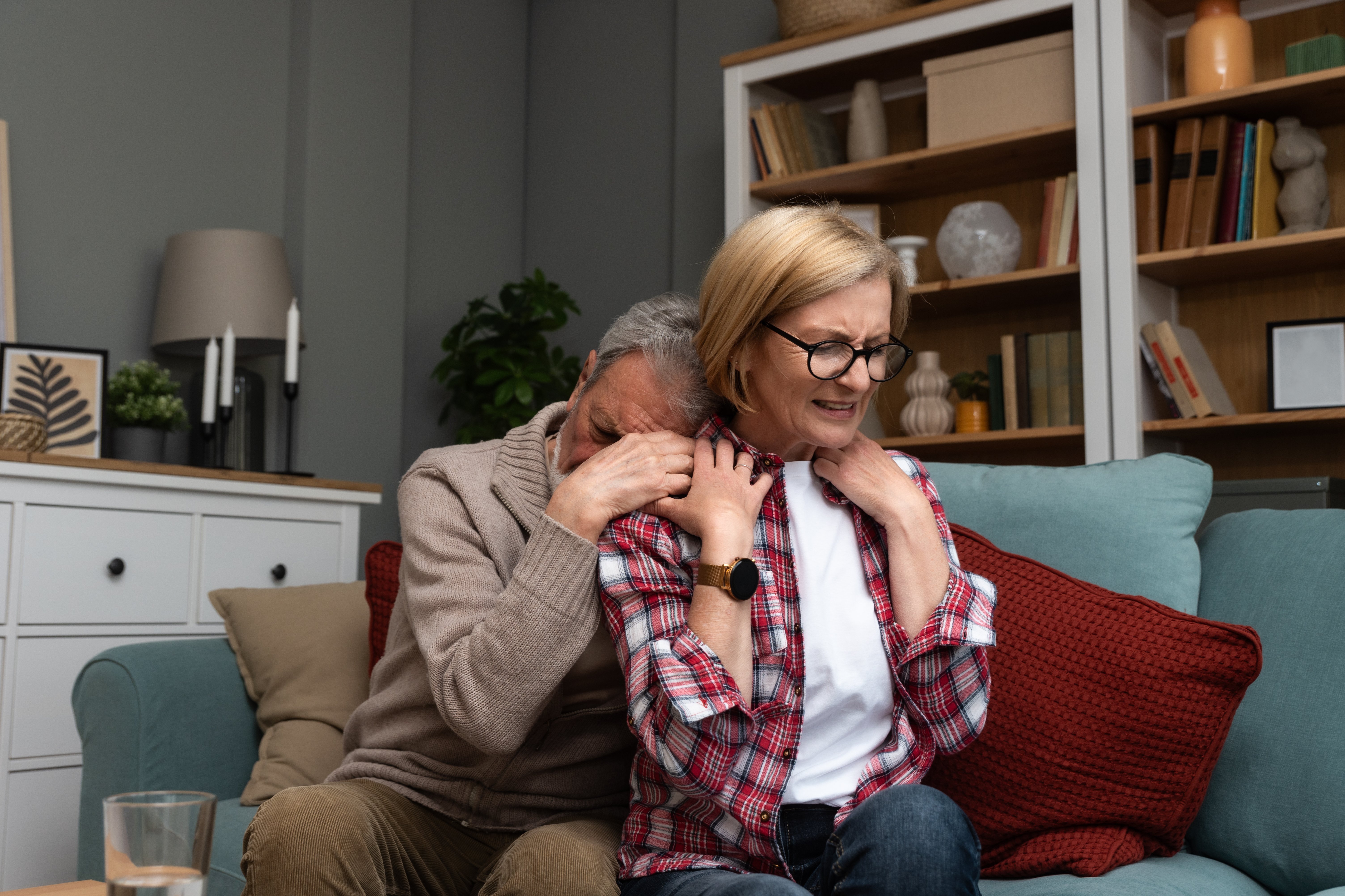 Anstrengende Angehörigenpflege Ein älterer Mann umarmt eine ältere Frau von hinten, sie sitzen auf einem Sofa. Er hat sein Gesicht von hinten in ihrer Schulter vergraben. Sie hält seine Hände, blickt geradeaus mit einem angestrengten, ernsten Ausdruck. Symbolfoto für Erschöpfung in der Angehörigenpflege.