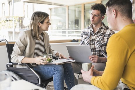 Inklusion am Arbeitsplatz Eine junge Frau im Rollstuhl spricht mit zwei Männern, wahrscheinlich Arbeitskollegen. Einer der Männer hat ein Notebook auf den Knien.