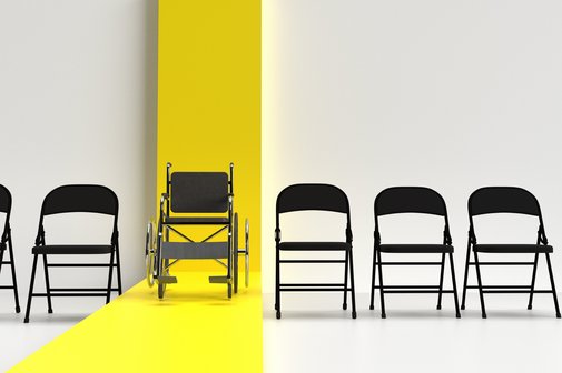 Klappstühle stehen in einer Reihe vor einer weißen Wand, in der Mitte ein Rollstuhl vor einem gelben Streifen, der sich über die Wand und den Fußboden zieht.