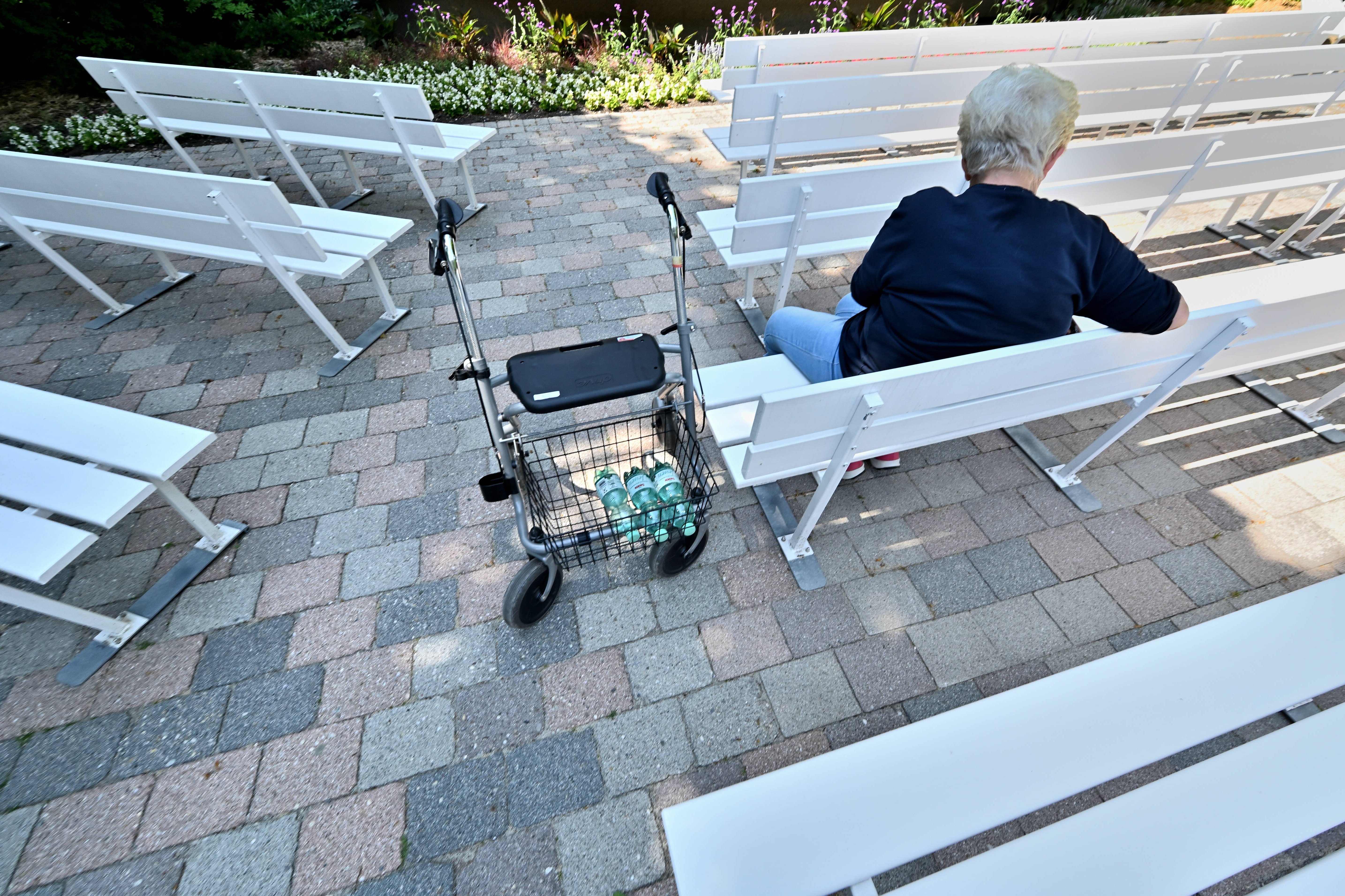 Eine ältere Dame sitzt alleine auf der Parkbank, neben ihr steht ein Rollator. 