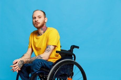 Ein junger Mann im gelben T-Shirt, der im Rollstuhl sitzt, vor blauem Hintergrund. Er blickt ernst in die Kamera.