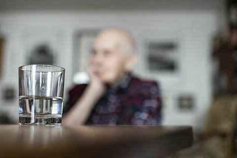 Einsamkeit bei Senioren Symbolfoto: Eine alte Frau sitzt allein an einem Tisch, den Kopf auf die Hand gestützt. Im Vordergrund steht ein Wasserglas auf dem Tisch.