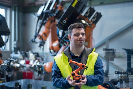 Ein junger Mann mit Downsyndrom in einer Werkshalle mit zahlreichen Maschinen, er trägt eine gelbe Sicherheitsweste
