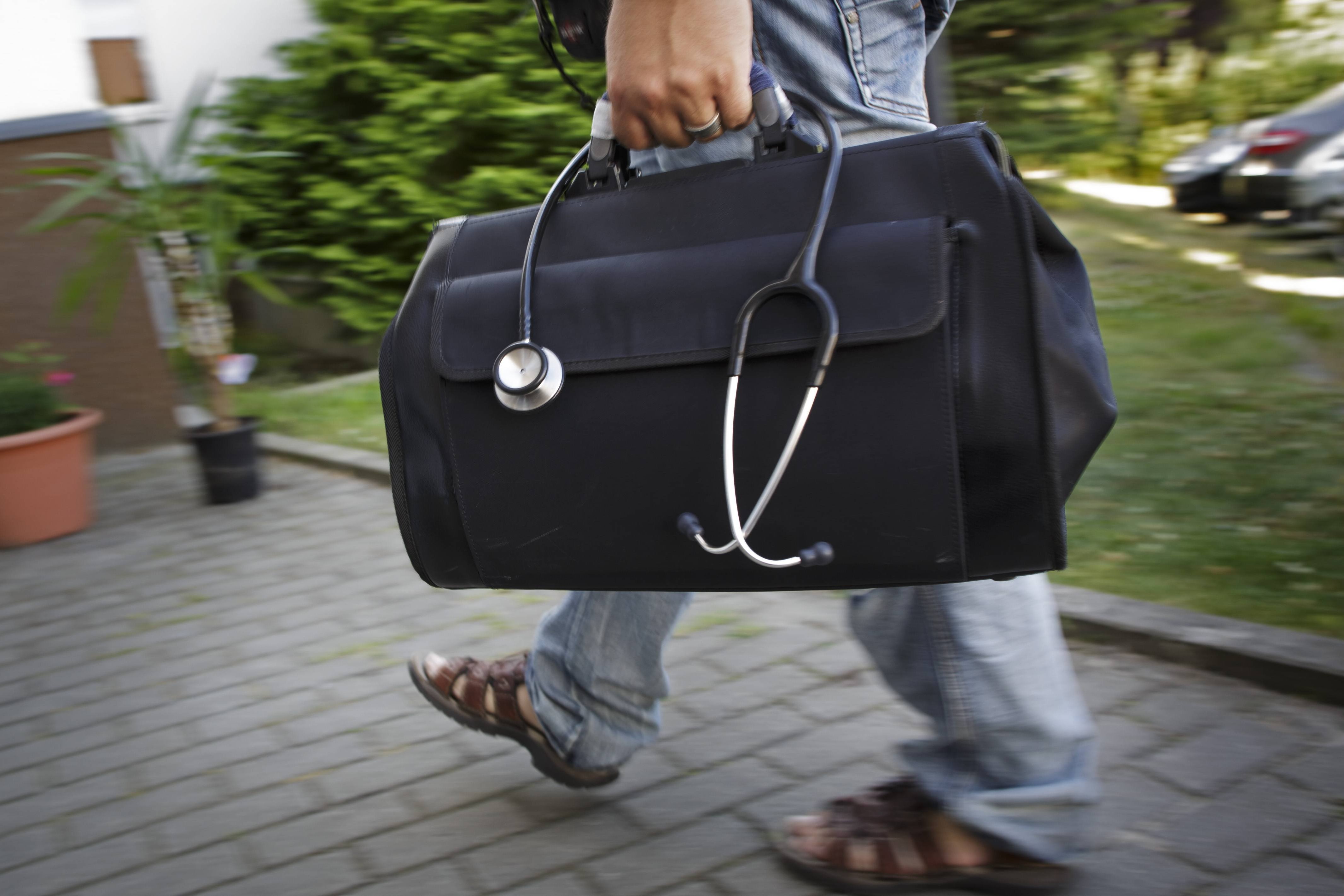 Ein Mann mit einer großen Arzttasche und einem Stethoskop geht einen gepflasterten Weg entlang. 