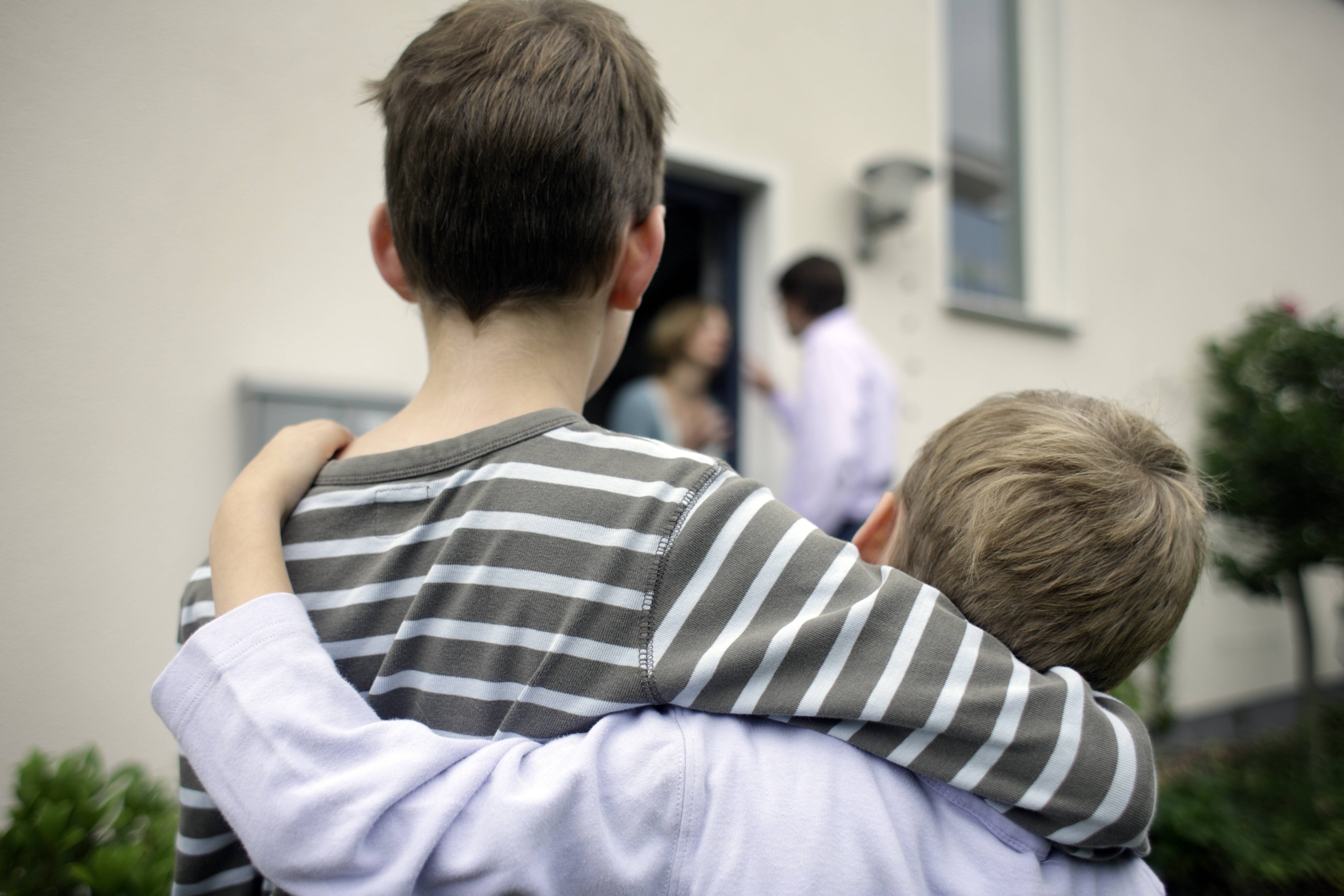 Zwei Jungen stehen Arm in Arm nebeneinander, im Hintergrund sieht man die Eltern streiten