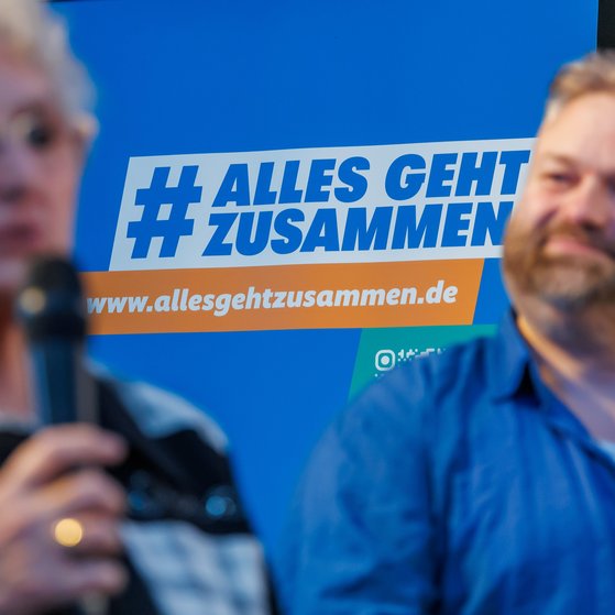 Zwei Ehrenamtliche beim Start der VdK-Aktion in Berlin: Eine Frau spricht in ein Mikrofon, ein Mann hört zu. Die beiden sind unscharf im Vordergrund zu sehen. Im Hintergrund sieht man scharf das Logo der Aktion Alles geht zusammen und den Link www.allesgehtzusammen.de