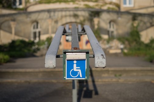 Eine Stufe mit einem Handlauf. An diesem ist eine Klingel mit einem Rollstuhl-Symbol angebracht; Symbol f&uuml;r eine nicht barrierefreie Stelle im &ouml;ffentlichen Raum.