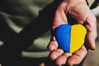 Hand einer älteren Person hält einen herzförmigen Stein, der zur Hälfte blau und zur Hälfte gelb angemalt ist in den Farben der ukrainischen Flagge