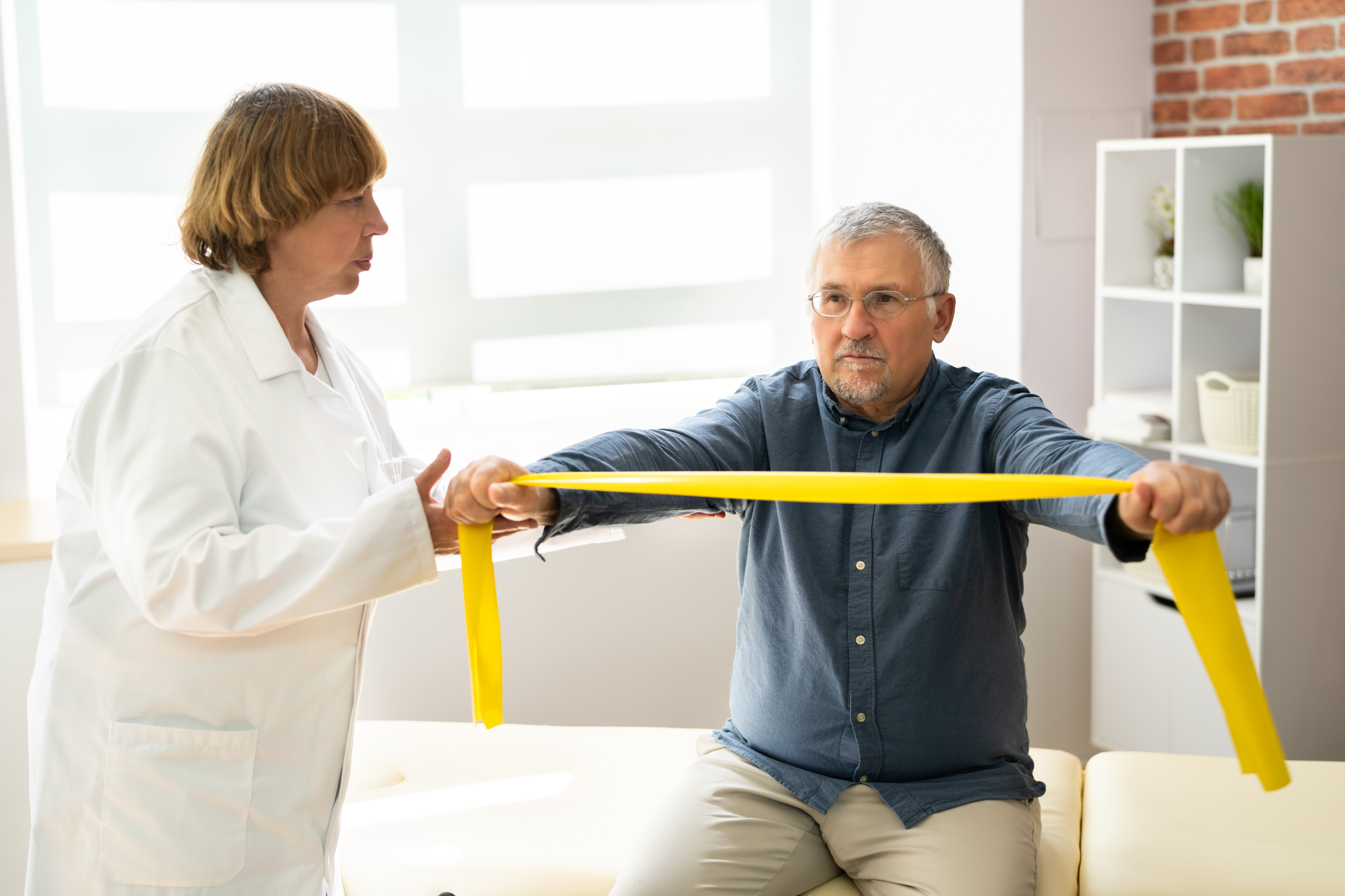 Geriatrische Reha Ein älterer Mann sitzt auf einer Liege in einer Praxis, er macht eine Übung mit einem gelben Thera-Band. Eine ältere Frau im weißen Kittel, die wie eine Physiotherapeutin aussieht, spricht mit ihm.